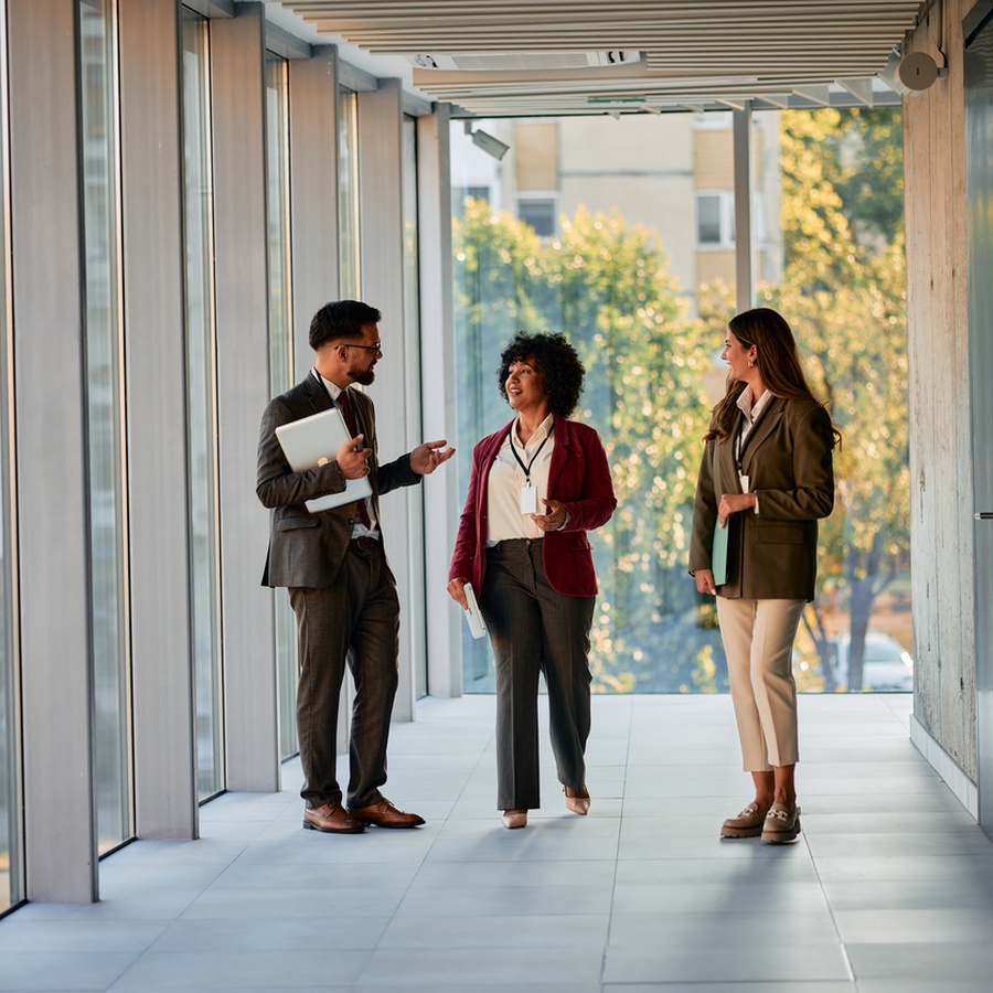 Three colleagues walking and talking in a glass-walled office corridor
